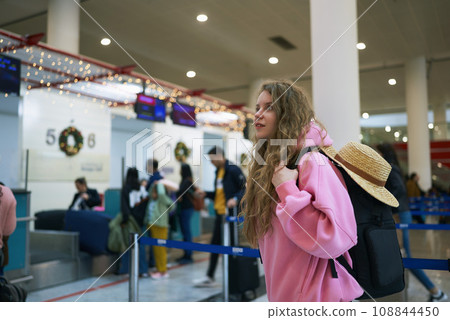Young female voyager at festive airport, adorned for Yuletide. Tourist awaits flight amid holiday rush, straw hat in hand, anticipation in her eyes. 108844450