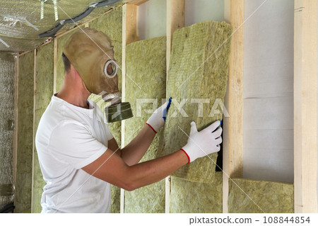 Young worker in gas mask insulating rock wool insulation staff in wooden frame for future house walls for cold and heat barrier. Comfortable warm home, economy, construction and renovation concept. Young worker in gas mask insulating rock wool insulation staff in wooden frame for future house walls for cold and heat barrier. Comfortable warm home, economy, construction and renovation concept. 108844854