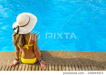 Young woman wearing yellow straw hat resting near swimming pool with clear blue water on summer sunny day. 108844855