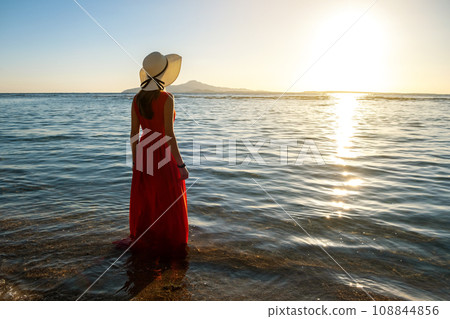Young woman wearing long red dress and straw hat standing in sea water at the beach enjoying view of rising sun in early summer morning. 108844856