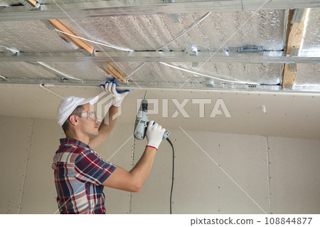 Young man in goggles fixing drywall suspended ceiling to metal frame using electrical screwdriver on ceiling insulated with shiny aluminum foil. Renovation, construction, do it yourself concept. 108844877