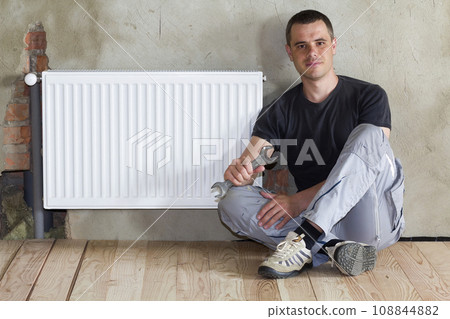 Young handsome plumber sitting on floor with wrench in hand near successfully installed heating radiator in empty room of a newly built apartment or house. Construction, maintenance and repair concept Young handsome plumber sitting on floor with wrench in hand near successfully installed heating radiator in empty room of a newly built apartment or house. Construction, maintenance and repair concept 108844882
