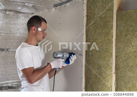 Worker in goggles with screwdriver working on insulation. Drywall on wall beams, insulating rock wool staff in wooden frame. Comfortable warm home, economy, construction and renovation concept. 108844898
