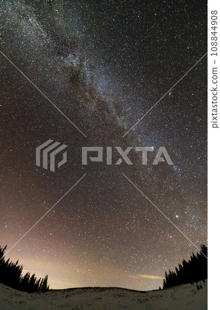 Winter mountains night landscape panorama. Milky Way bright constellation in dark blue starry sky over dark spruce pine trees forest, soft glow on horizon after sunset. Wide angle shot. 108844908