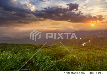 Wide summer mountain view at sunrise. Glowing orange sun raising in blue cloudy sky over green grassy hill soft grass and distant mountain range covered with morning mist. Beauty of nature concept. Wide summer mountain view at sunrise. Glowing orange sun raising in blue cloudy sky over green grassy hill soft grass and distant mountain range covered with morning mist. Beauty of nature concept. 108844939