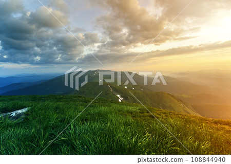Wide summer mountain view at sunrise. Glowing orange sun raising in blue cloudy sky over green grassy hill soft grass and distant mountain range covered with morning mist. Beauty of nature concept. Wide summer mountain view at sunrise. Glowing orange sun raising in blue cloudy sky over green grassy hill soft grass and distant mountain range covered with morning mist. Beauty of nature concept. 108844940