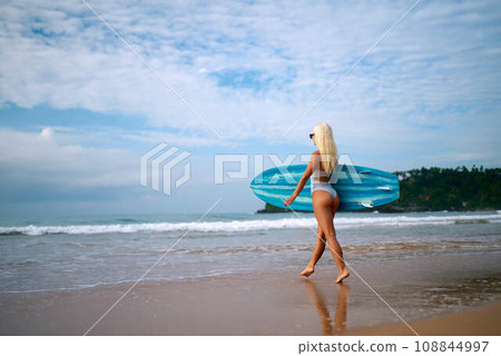 Blonde woman in white bikini holding blue surfboard on sandy beach, ready to surf on ocean waves, tropical coastline with greenery in background, active summer water sports, female surfer lifestyle. Blonde woman in white bikini holding blue surfboard on sandy beach, ready to surf on ocean waves, tropical coastline with greenery in background, active summer water sports, female surfer lifestyle. 108844997