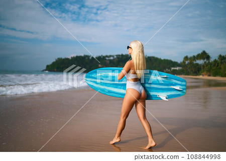 Blonde woman in white bikini carries blue surfboard on tropical beach, ready for surfing lesson. Tourist enjoys summer vacation, beach lifestyle on sunny coastline. Blonde woman in white bikini carries blue surfboard on tropical beach, ready for surfing lesson. Tourist enjoys summer vacation, beach lifestyle on sunny coastline. 108844998