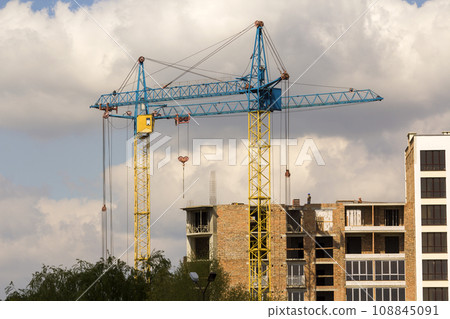 Urban view of silhouettes of two high industrial tower cranes above green tree tops working at construction of new brick building with workers in hard hats on it against bright blue sky background. Urban view of silhouettes of two high industrial tower cranes above green tree tops working at construction of new brick building with workers in hard hats on it against bright blue sky background. 108845091
