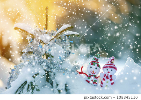 Two small funny toys baby snowman in knitted hats and scarves in deep snow outdoors near pine tree branch. Happy New Year and Merry Christmas greeting card. 108845110