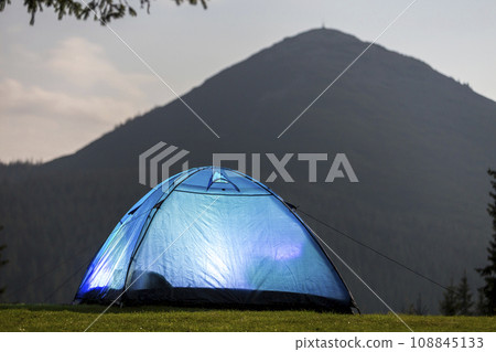 Tourist hikers bright blue tent on green grassy forest clearing among tall pine trees under clear morning sky on distant foggy woody mountain background. Tourism, summer camping in mountains at dawn. 108845133