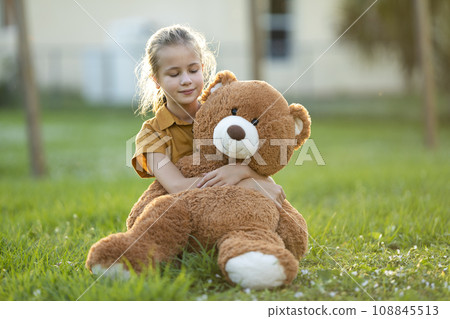 Pretty child girl tenderly embracing her teddy bear friend outdoors on green grass lawn. Concept of friendship values Pretty child girl tenderly embracing her teddy bear friend outdoors on green grass lawn. Concept of friendship values 108845513