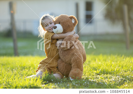 Pretty child girl tenderly embracing her teddy bear friend outdoors on green grass lawn. Concept of friendship values Pretty child girl tenderly embracing her teddy bear friend outdoors on green grass lawn. Concept of friendship values 108845514