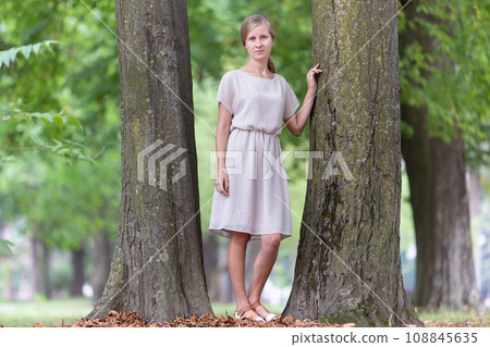 Portrait of a young woman standing near big tree trunk in summer park outdoors. 108845635