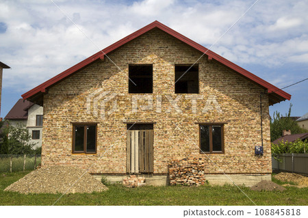 New not finished brick house with boarded door entrance, plastic windows on first floor, piles of brick and gravel on green grass on fenced land plot. Construction, investment and real property. 108845818