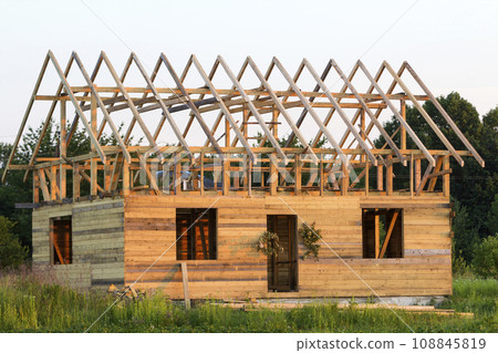 New cottage of natural ecological lumber materials under construction in green field. Wooden walls and steep roof frame. Property, investment, professional building and reconstruction concept. 108845819