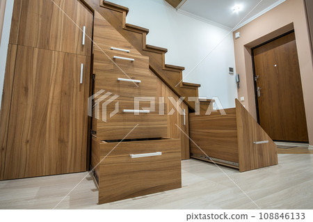 Modern architecture interior with luxury hallway with glossy wooden stairs in multi-storey house. Custom built pullout cabinets on glides in slots under stairs. Use of space for storage. Modern architecture interior with luxury hallway with glossy wooden stairs in multi-storey house. Custom built pullout cabinets on glides in slots under stairs. Use of space for storage. 108846133