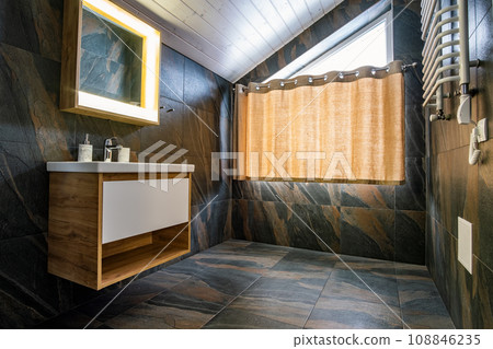 Interior of modern stylish bathroom with black tiled walls, curtain shower place and wooden furniture with wash basin and big illuminated mirror. 108846235