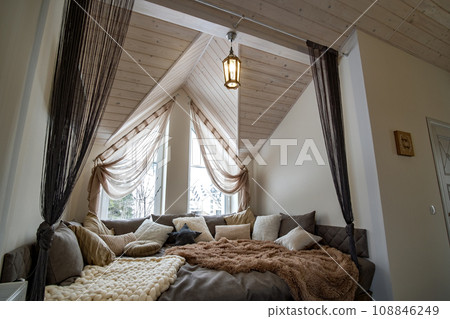 Interior of a modern house spacious hallway with big soft resting place. Contemporary wide sofa with many pillows and light window onder wooden loft ceiling. 108846249