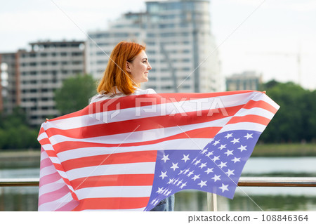 Happy young red haired woman with USA national flag on her shoulders standing outdoors in american city. Positive girl celebrating United States independence day. Happy young red haired woman with USA national flag on her shoulders standing outdoors in american city. Positive girl celebrating United States independence day. 108846364
