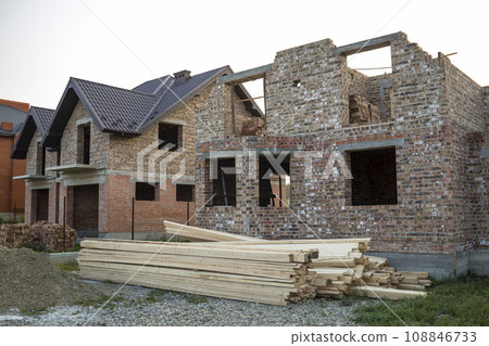Future cottage under construction and pile of gravel and stack of boards in front of not finished new big brick house with brown shingled roof. Construction, investment and real property concept. 108846733