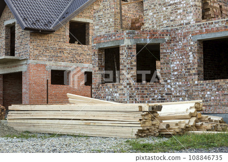 Future cottage under construction and pile of gravel and stack of boards in front of not finished new big brick house with brown shingled roof. Construction, investment and real property concept. Future cottage under construction and pile of gravel and stack of boards in front of not finished new big brick house with brown shingled roof. Construction, investment and real property concept. 108846735