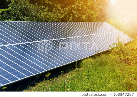 Close-up surface of lit by sun modern saving efficient stand -alone blue shiny solar photo voltaic panels system producing renewable clean energy on green grass and trees foliage background. Close-up surface of lit by sun modern saving efficient stand -alone blue shiny solar photo voltaic panels system producing renewable clean energy on green grass and trees foliage background. 108847297