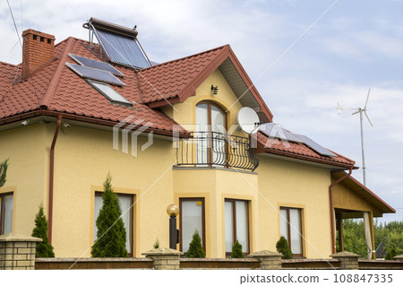 Close-up of new brick house top with red shingle roof, satellite dish, solar panels and plastic attic windows on bright blue sky background. Real estate property and professionally done work concept. Close-up of new brick house top with red shingle roof, satellite dish, solar panels and plastic attic windows on bright blue sky background. Real estate property and professionally done work concept. 108847335