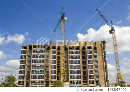 Building site with two industrial tower cranes working at construction of new brick tall building on bright blue sky and green trees background. 108847401
