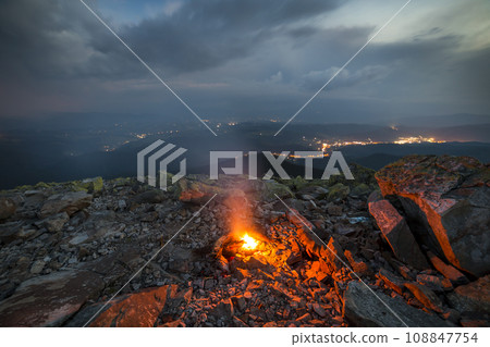 Beautiful summer night in mountain. Bright fire burns on rocky mountain top under blue cloudy sky. Beautiful summer night in mountain. Bright fire burns on rocky mountain top under blue cloudy sky. 108847754