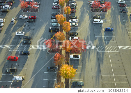 Aerial view of many colorful cars parked on parking lot with lines and markings for parking places and directions. Place for vehicles in front of a strip mall plaza Aerial view of many colorful cars parked on parking lot with lines and markings for parking places and directions. Place for vehicles in front of a strip mall plaza 108848214