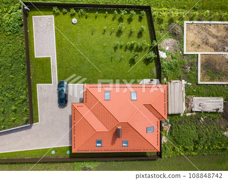 Aerial top view of house shingle roof with attic windows and black car on paved yard with green grass lawn. 108848742