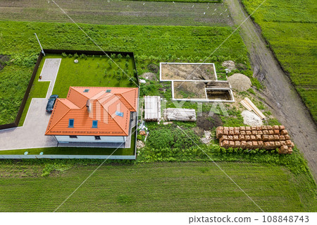 Aerial top view of a private house with attic windows on roof, paved yard with green grass lawn and building site with concrete foundation floor and stacks of yellow bricks for construction.. Aerial top view of a private house with attic windows on roof, paved yard with green grass lawn and building site with concrete foundation floor and stacks of yellow bricks for construction.. 108848743