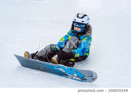little boy sitting on snow putting his feet in snowboard bindings adjusting straps. little boy sitting on snow putting his feet in snowboard bindings adjusting straps. 108850349
