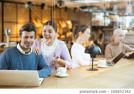 Portrait of middle-aged male and young female surfing Internet on notebook while drinking tea or coffee in cafeteria Portrait of middle-aged male and young female surfing Internet on notebook while drinking tea or coffee in cafeteria 108852162