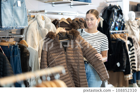 Girl stopped near showcase with outerwear, holds fur coat hanger in hands and examines product Girl stopped near showcase with outerwear, holds fur coat hanger in hands and examines product 108852310