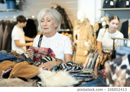 Senior woman buyer carefully sort through various wardrobe items presented in window of second-hand store Senior woman buyer carefully sort through various wardrobe items presented in window of second-hand store 108852413