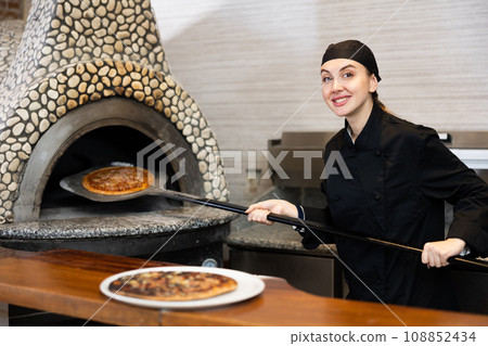 Portrait of female chef in black uniform holding big shovel with pizza in kitchen 108852434