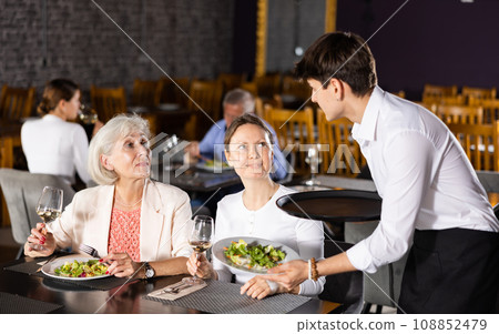 Young male waiter brought food to order of young girl and elderly mother to visitors of restaurant. Young male waiter brought food to order of young girl and elderly mother to visitors of restaurant. 108852479