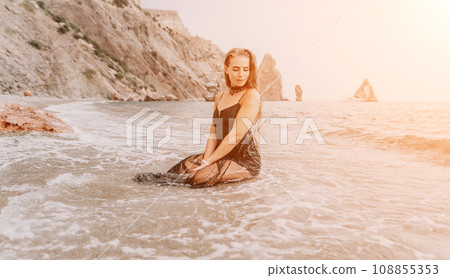 Woman summer travel sea. Happy tourist in black dress enjoy taking picture outdoors for memories. Woman traveler posing on sea beach surrounded by volcanic mountains, sharing travel adventure journey 108855353