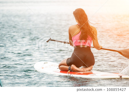 Sea woman sup. Silhouette of happy young woman in pink bikini, surfing on SUP board, confident paddling through water surface. Idyllic sunset. Active lifestyle at sea or river. 108855427