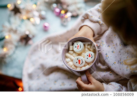 Unrecognizable little child girl holding cup with hot chocolate with marshmallows as snowman. Kid sitting near Christmas decorated window with lights. Unrecognizable little child girl holding cup with hot chocolate with marshmallows as snowman. Kid sitting near Christmas decorated window with lights. 108856009