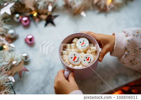 Unrecognizable little child girl holding cup with hot chocolate with marshmallows as snowman. Kid sitting near Christmas decorated window with lights. 108856011