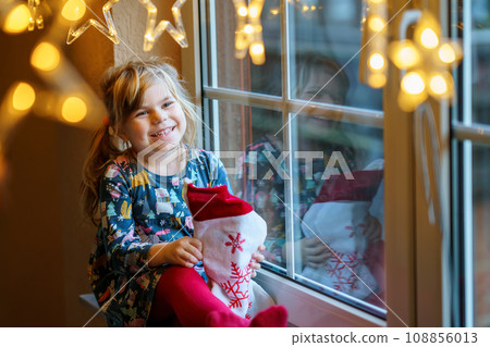 Little preschool girl holding cup Santa Claus boot with gift called Nikolausstiefel in German. Happy child wait on holiday by window with Christmas lights in winter. Cozy family celebration of xmas. 108856013