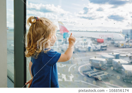 Cute little toddler girl at the airport, traveling. Happy healthy child waiting near window and watching airplanes. Family going on summer vacations by plane. 108856174