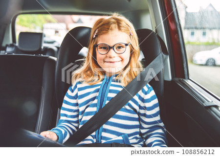 Little Preschool Girl Sitting in Her Car Seat. Happy Child with Eyeglasses Reading a Book, Smiling on the Way to Family Vacations during Traffic Jam. 108856212