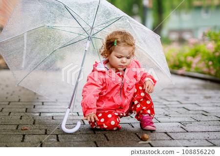 Cute adorable baby girl discovering snail on a walk. Beautiful curly toddler child having fun on rainy day. With big umbrella, child in waterproof clothes Cute adorable baby girl discovering snail on a walk. Beautiful curly toddler child having fun on rainy day. With big umbrella, child in waterproof clothes 108856220