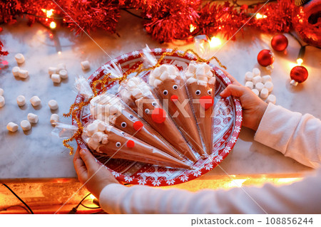 Close-up of little girl holding reindeer made with chocolate cocoa powder and marshmallows. Selfmade dessert drink for children on Christmas. Closeup of hands of child 108856244