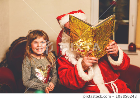 Little toddler girl talking to Santa Claus called Nikolaus or Weihnachtsmann in German. Happy smiling kid waiting for gifts. Santa with golden book. Xmas, childhood, holiday Little toddler girl talking to Santa Claus called Nikolaus or Weihnachtsmann in German. Happy smiling kid waiting for gifts. Santa with golden book. Xmas, childhood, holiday 108856248