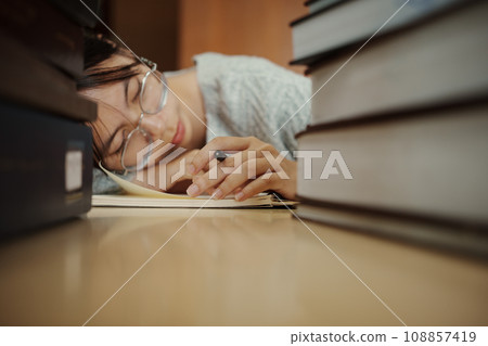 Tired student girl with glasses lying on books in library. 108857419